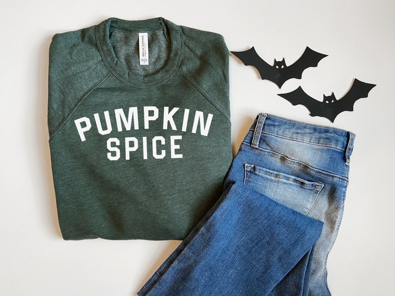 May include: A green sweatshirt with the text "PUMPKIN SPICE" in white letters. The sweatshirt is folded and laid on a white surface next to a pair of blue jeans and two black paper bats.