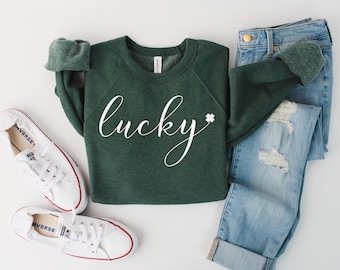 May include: A green sweatshirt with the word "lucky" written in white cursive script with a four-leaf clover next to it. The sweatshirt is laid out on a white surface with a pair of white sneakers and blue jeans.