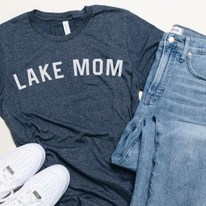 Op de afbeelding: Een casual outfit met een donkergrijs t-shirt met de tekst "LAKE MOM" in het wit, gecombineerd met lichtblauwe jeans en witte sneakers. De set is op een wit oppervlak geplaatst, wat wijst op een lifestyle- of modethema.