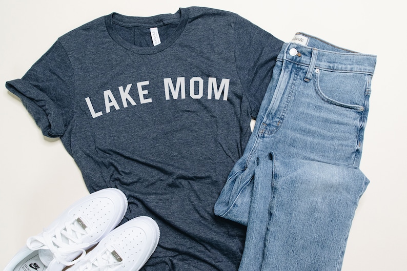 Op de afbeelding: Een casual outfit met een donkergrijs t-shirt met de tekst "LAKE MOM" in het wit, gecombineerd met lichtblauwe jeans en witte sneakers. De set is op een wit oppervlak geplaatst, wat wijst op een lifestyle- of modethema.