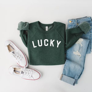 May include: A green sweatshirt with the word "LUCKY" printed in white on the front. The sweatshirt is paired with blue jeans and white sneakers.