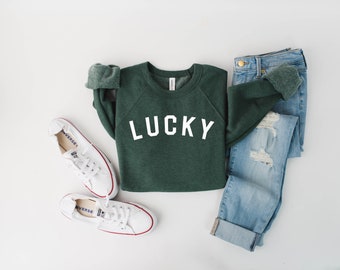 May include: A green sweatshirt with the word "LUCKY" printed in white on the front. The sweatshirt is paired with blue jeans and white sneakers.