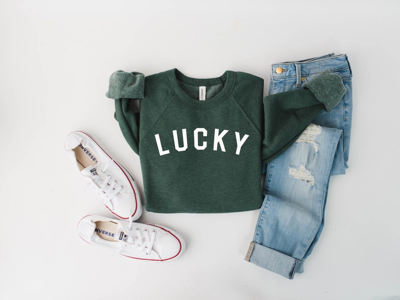 May include: A green sweatshirt with the word "LUCKY" printed in white on the front. The sweatshirt is paired with blue jeans and white sneakers.