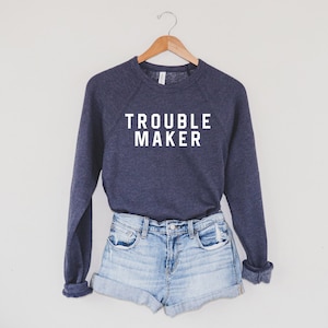 May include: A navy blue sweatshirt with the text "TROUBLE MAKER" in white block letters. The sweatshirt is displayed with a pair of light blue denim shorts on a wooden hanger against a white background.