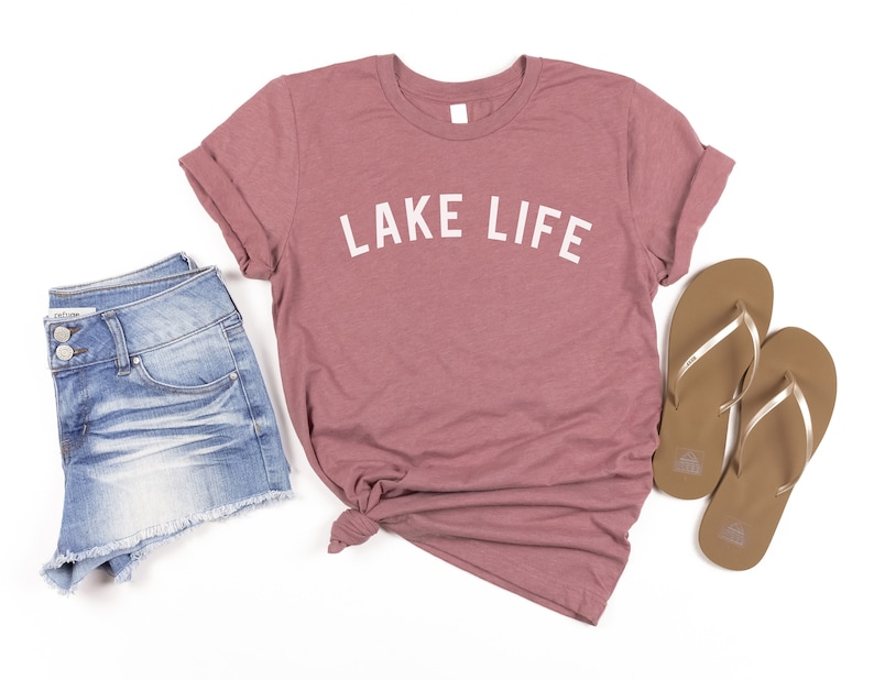 May include: A mauve t-shirt with the text "LAKE LIFE" in white lettering. The t-shirt is paired with denim shorts and brown flip-flops.