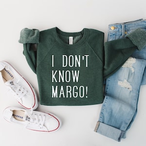 May include: A green sweatshirt with the text "I don't know Margo!" in white lettering. The sweatshirt is laid out on a white surface with a pair of white sneakers and a pair of blue jeans.
