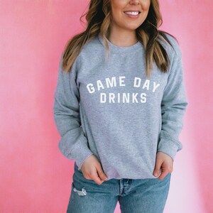 K&ouml;nnte beinhalten: Ein grauer Pullover mit dem Text "GAME DAY DRINKS" in wei&szlig;en Buchstaben.