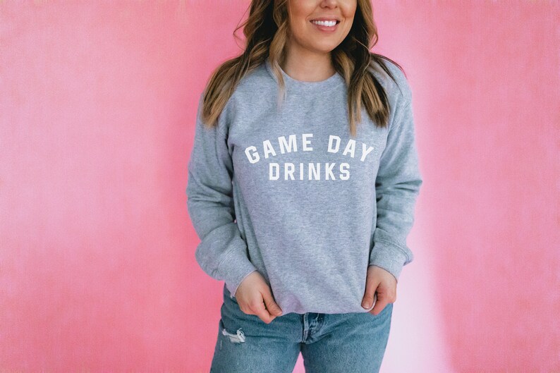 K&ouml;nnte beinhalten: Ein grauer Pullover mit dem Text "GAME DAY DRINKS" in wei&szlig;en Buchstaben.