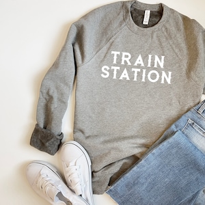 Könnte beinhalten: Ein grauer Pullover mit dem Aufdruck "TRAIN STATION" in Weiß auf der Vorderseite. Der Pullover wird mit einer blauen Jeans und weißen Turnschuhen kombiniert.
