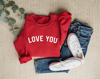 May include: Red sweatshirt with white text that says "LOVE YOU" on a light brown surface with blue jeans and white sneakers.
