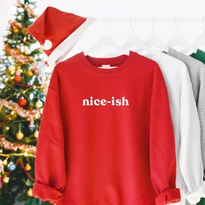 May include: A red crew neck sweatshirt with the text "nice-ish" in white. Other sweatshirts in white, grey, and green are on a rail. A Santa hat is above the red sweatshirt. A decorated Christmas tree is in the background.