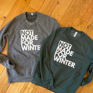 May include: Two crewneck sweatshirts, one dark gray and one dark green, with the text "NOT MADE FOR WINTER" in white block letters. The sweatshirts are displayed on a wooden surface.