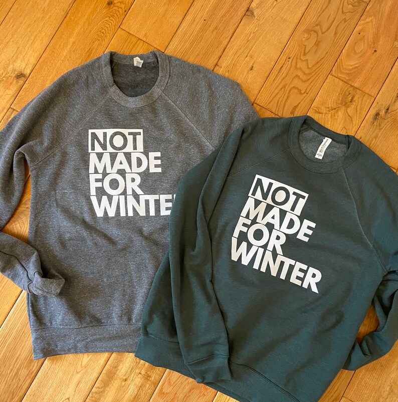 May include: Two crewneck sweatshirts, one dark gray and one dark green, with the text "NOT MADE FOR WINTER" in white block letters. The sweatshirts are displayed on a wooden surface.