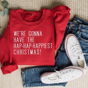 May include: A red sweatshirt with the text "WE'RE GONNA HAVE THE HAP-HAP-HAPPIEST CHRISTMAS!" in white. The sweatshirt is paired with blue jeans and white sneakers with red accents, creating a casual holiday outfit.