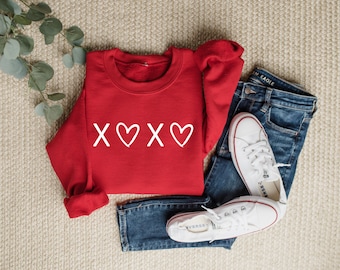 May include: Red sweatshirt with white 'XOXO' graphic with hearts. The sweatshirt is laid out on a beige surface with a pair of blue jeans and white sneakers.