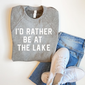 May include: A gray sweatshirt with the text "I'd rather be at the lake" in white lettering. The sweatshirt is folded and laid on a white surface with a pair of blue jeans and white sneakers.