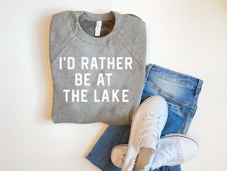 May include: A gray sweatshirt with the text "I'd rather be at the lake" in white lettering. The sweatshirt is folded and laid on a white surface with a pair of blue jeans and white sneakers.