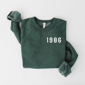 May include: A dark green sweatshirt with the year "1986" printed in white block letters. The long-sleeved top is laid flat on a white surface, with the sleeves folded. The crew neck sweatshirt is made of a soft material.