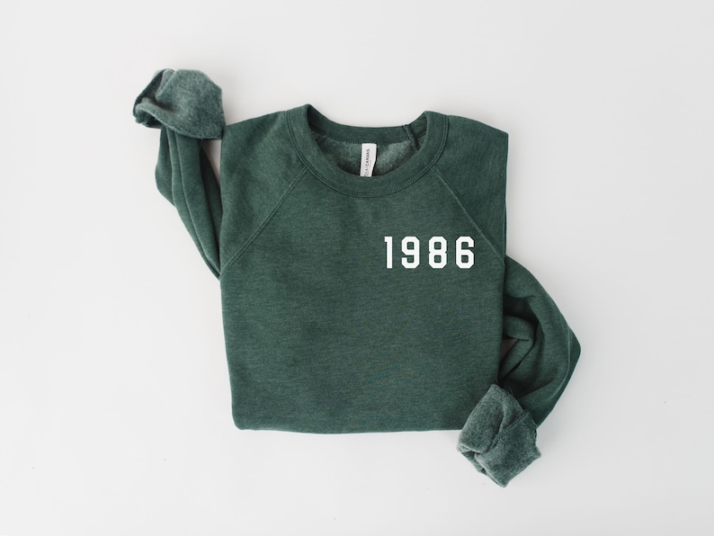 May include: A dark green sweatshirt with the year "1986" printed in white block letters. The long-sleeved top is laid flat on a white surface, with the sleeves folded. The crew neck sweatshirt is made of a soft material.