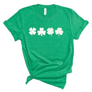 May include: A green t-shirt with a white clover design. The shirt is a solid green color and features four white shamrock designs across the chest. The shirt is made of a soft material and has short sleeves.