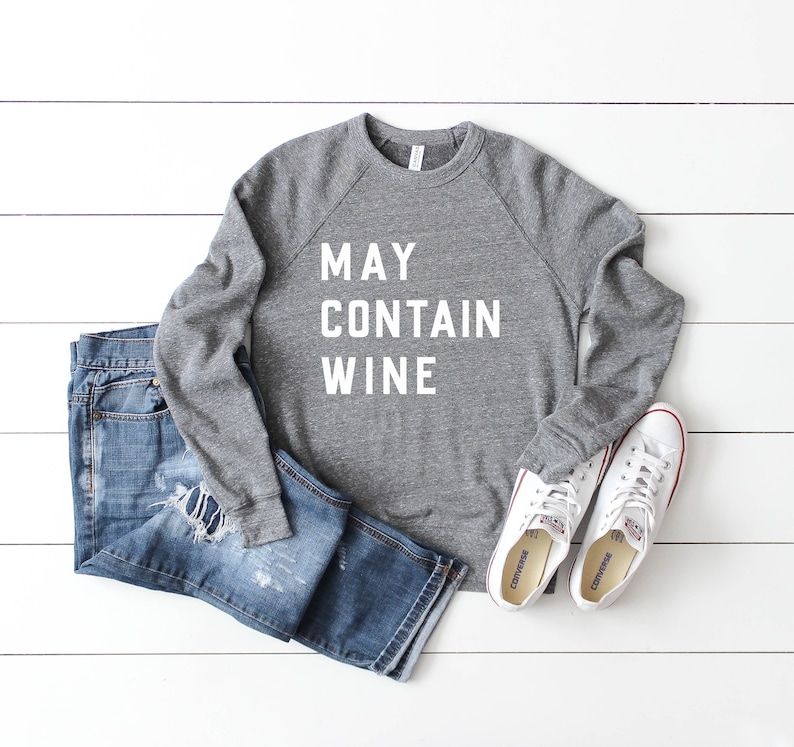 Puede incluir: Sudadera gris con el texto "MAY CONTAIN WINE" en letras blancas. La sudadera se combina con jeans azules y zapatillas blancas.