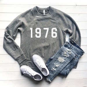 Può includere: Una felpa grigia con il numero "1976" stampato in bianco, abbinata a jeans blu strappati e scarpe da ginnastica bianche. L'outfit casual è disposto su una superficie di legno bianco, creando uno stile semplice e quotidiano.