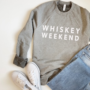 May include: A gray sweatshirt with the text "Whiskey Weekend" printed in white on the front. The sweatshirt is laid out on a white surface with a pair of white sneakers and blue jeans.