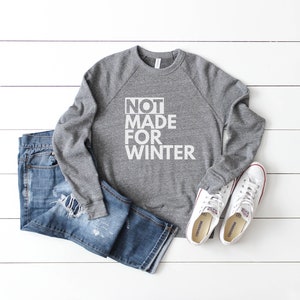 May include: A gray sweatshirt with the text "NOT MADE FOR WINTER" in white, paired with distressed blue jeans and white canvas sneakers with red accents. The items are arranged on a white wooden surface.