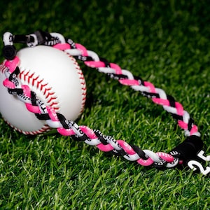 Op de afbeelding: Een witte honkbal met rode stiksels wordt gedeeltelijk omcirkeld door een gevlochten ketting. De ketting heeft roze, zwarte en witte strengen. Een zwarte sluiting en een witte bedel met nummer 25 zijn ook zichtbaar.