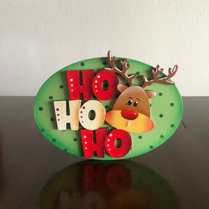 May include: A green oval wooden sign with white polka dots and the words "HO HO HO" in red and white letters. A cartoon reindeer face is in the center of the sign.