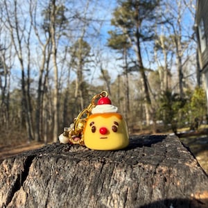 May include: A yellow pudding charm with a cartoon face, topped with white cream and a red cherry. The charm is attached to a gold keychain with beads. The charm is sitting on a weathered tree stump.