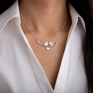 Può includere: Collana d'argento con due ciondoli a forma di uccello su un ramo curvo e un piccolo cuore. La collana è indossata su una camicia bianca.