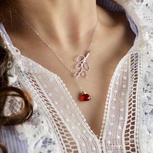Puede incluir: Un delicado collar de plata con un diseño de hoja y una gema roja colgante. El collar se lleva sobre una blusa blanca con encaje y una camisa a rayas azules. La joya añade un toque de elegancia.