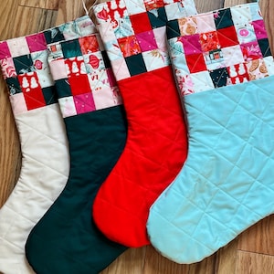 Puede incluir: Cuatro calcetines navideños acolchados en blanco, verde oscuro, rojo y azul claro. Cada calcetín tiene un cuerpo acolchado y un puño de patchwork con patrones festivos en rojo, rosa y verde. Se muestran sobre una superficie de madera.