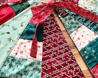May include: A patchwork Christmas tree skirt with a variety of festive patterns in red, green, and white. The skirt features a central opening and is tied with a red ribbon. The fabrics include holiday motifs like mittens, snowflakes, and trees.