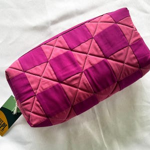 Puede incluir: Un neceser acolchado en tonos rosa y magenta. La bolsa presenta un patrón geométrico de cuadrados y diamantes. Una pequeña etiqueta con texto y una borla está unida a la cremallera.