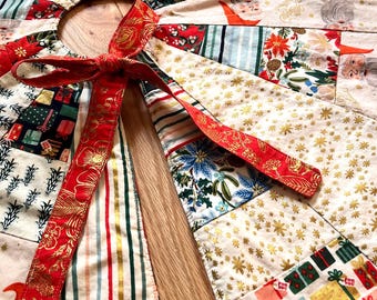May include: A patchwork Christmas tree skirt with a variety of festive patterns. The skirt features gift boxes, floral designs, and gold star accents. A red ribbon with gold floral detailing is tied at the opening. The skirt is designed to wrap around a tree base.