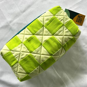 May include: A quilted cosmetic bag with a geometric pattern of lime green squares on a light green background. The bag has a teal zipper and a tag that reads "WEST MORE PL LAND".