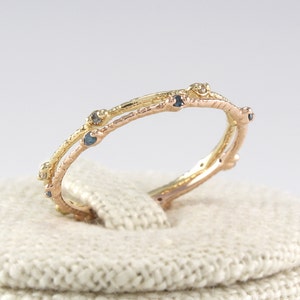 Könnte beinhalten: Ein zarter, zweifarbiger Ring mit einem astähnlichen Design. Der Ring hat eine Kombination aus Gold und Roségold, verziert mit kleinen, dunkelblauen Edelsteinen. Der Ring wird auf einer weißen, strukturierten Oberfläche präsentiert.