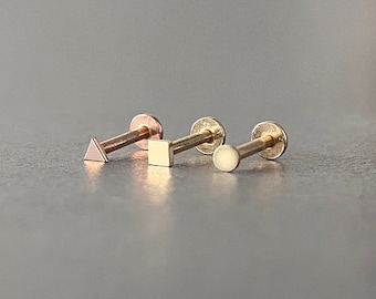 May include: Three gold body jewelry pieces with different geometric shapes: a triangle, a square, and a circle. The jewelry is on a gray surface.