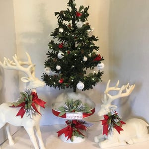 May include: A small artificial Christmas tree decorated with white ornaments, red cardinal figures, and lights, flanked by two white deer figurines. The tree sits on a white pedestal stand with a red ribbon and a sign that reads CHRISTMAS.