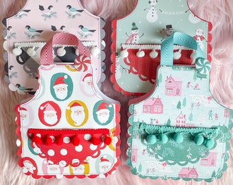 Könnte beinhalten: Vier festliche Weihnachts-Schürzen mit verschiedenen Designs. Jede Schürze hat einen gewellten Rand und eine Tasche mit Bommeln. Die Designs beinhalten einen Schneemann, den Weihnachtsmann und ein Winterdorf.
