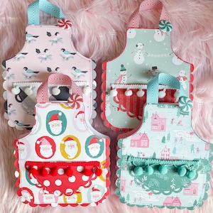 Könnte beinhalten: Vier festliche Weihnachts-Schürzen mit verschiedenen Designs. Jede Schürze hat einen gewellten Rand und eine Tasche mit Bommeln. Die Designs beinhalten einen Schneemann, den Weihnachtsmann und ein Winterdorf.