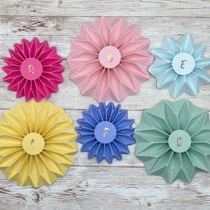 Può includere: Sei decorazioni di fiori di carta in vari colori, tra cui rosa, giallo, blu e verde. Ogni fiore ha una lettera al centro, che compone la parola "ABCDE".