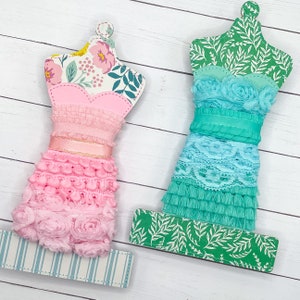 May include: Two fabric covered dress forms with different lace and trim embellishments. The dress form on the left is pink with floral print and the dress form on the right is turquoise with a green floral print.