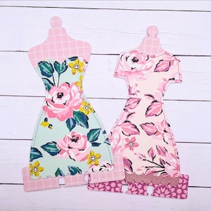 Könnte beinhalten: Zwei Papierpuppen-Kleiderformen mit Blumenmustern. Das Kleid links ist grün mit rosa und gelben Blumen. Das Kleid rechts ist rosa mit weißen und rosa Blumen.