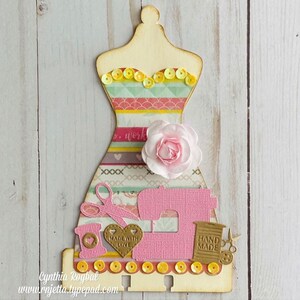 K&ouml;nnte beinhalten: Eine h&ouml;lzerne Kleiderpuppe mit einem rosa-gelb gestreiften Kleid, einer rosa N&auml;hmaschine, einer Garnrolle, einer Schere und einem Herz mit dem Text "Made with Love".