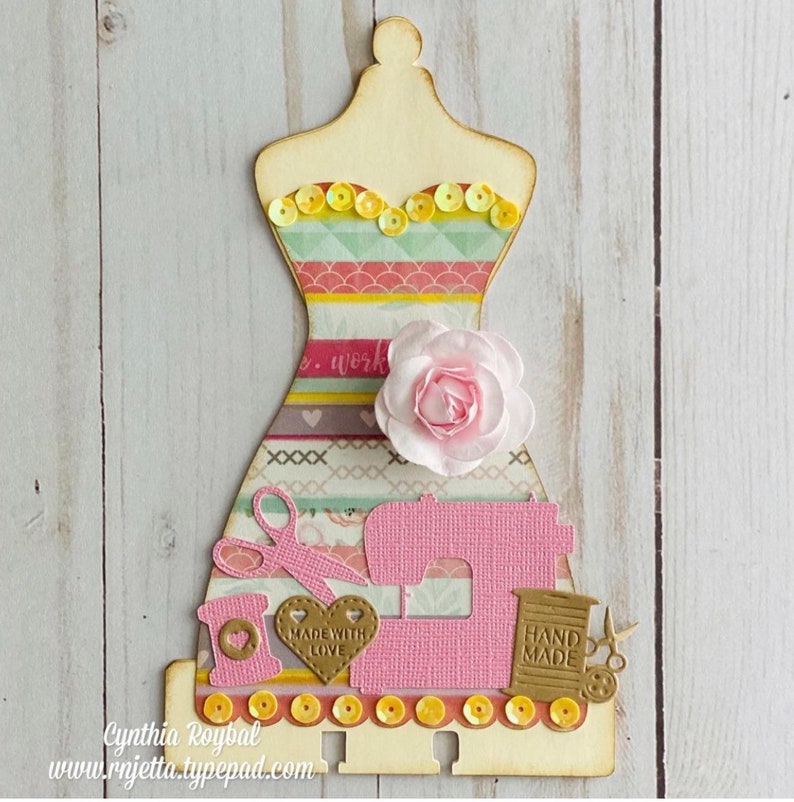 K&ouml;nnte beinhalten: Eine h&ouml;lzerne Kleiderpuppe mit einem rosa-gelb gestreiften Kleid, einer rosa N&auml;hmaschine, einer Garnrolle, einer Schere und einem Herz mit dem Text "Made with Love".