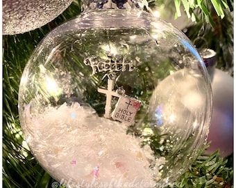 May include: Clear glass Christmas ornament with a silver metal cross, a tiny Holy Bible charm, and the word "Faith" at the top. The ornament is filled with white, sparkly material and is hanging on a Christmas tree.