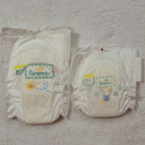 May include: Two white Pampers diapers, one larger than the other, are displayed. The diapers have a green and yellow design with the Pampers logo and a bee illustration. One diaper has a pin in it.